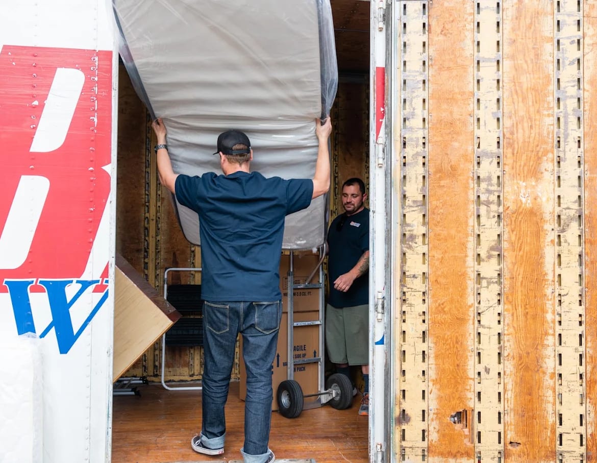 A BEST MOVE mover lifting a bagged mattress into the back of a lettered BEST MOVE truck while a second mover steadies the load