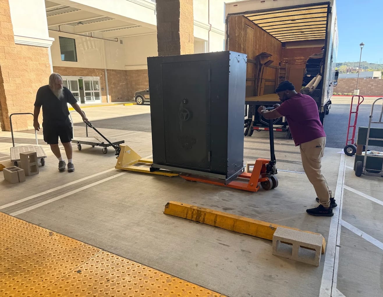 Two BEST MOVE movers walking a tall steel gun safe across a loading dock on a pallet jack