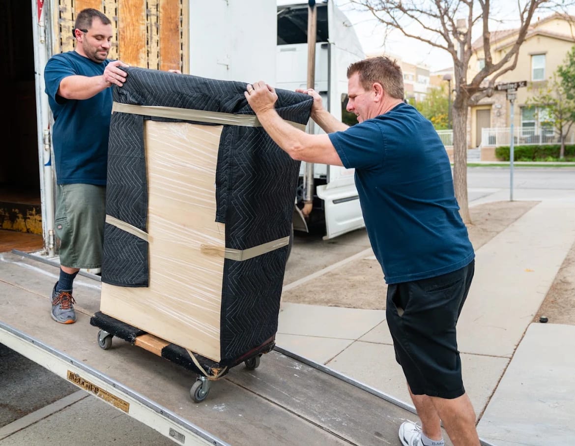 Two BEST MOVE movers wheeling a pad-wrapped and shrink-wrapped chest of drawers up the truck ramp on a four-wheel dolly