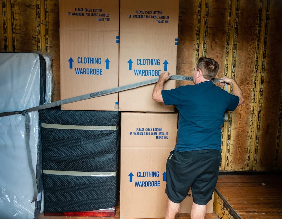 A BEST MOVE mover pulling a strap tight across a stacked tier of clothing wardrobe boxes inside the truck