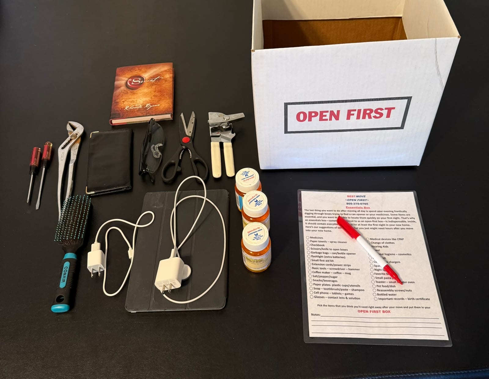 The bright white Open First Box carton on a black background, beside its essentials checklist and marker, with first-night items laid out: tools, a can opener, scissors, phone chargers, medicines, a hairbrush and a book
