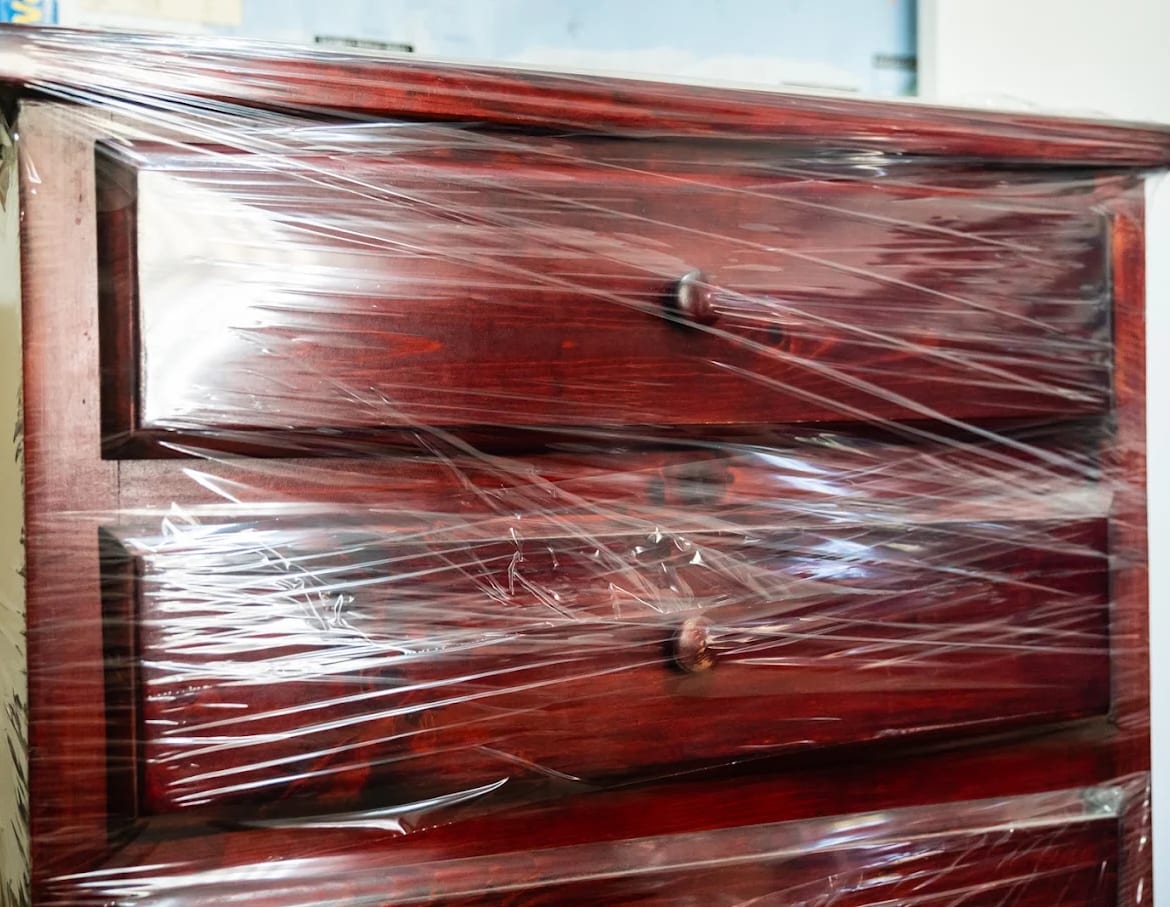A wooden chest of drawers shrink-wrapped for transit, with the drawers held shut